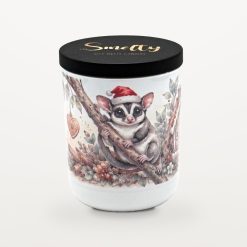 Smelty Christmas candle in a white ceramic jar with a black wooden lid and gold Smelty logo, printed with a big-eyed sugar glider in a Santa hat clinging to a gum branch beside a hanging gingerbread heart ornament.